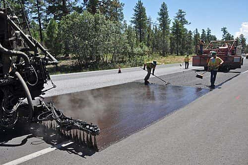 A truck-mounted distributor sprays hot liquid asphalt onto a highway lane while crew members sweep behind it