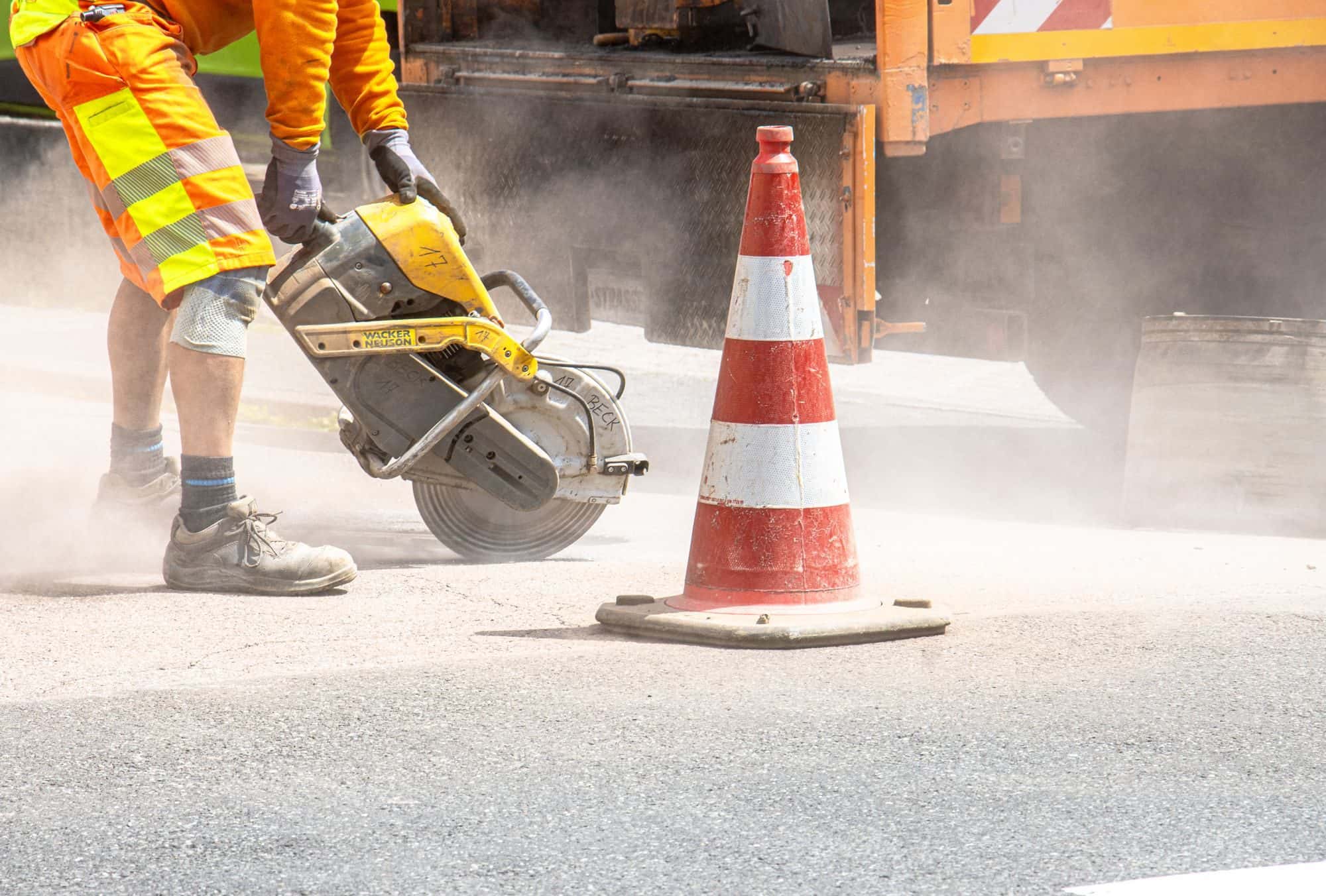 A worker cuts pavement with a gas-powered saw next to a traffic cone as dust rises