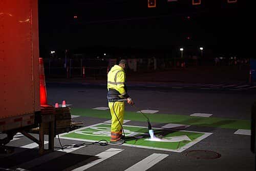 A worker in high-visibility rain gear heats a thermoplastic pavement marking with a torch at night