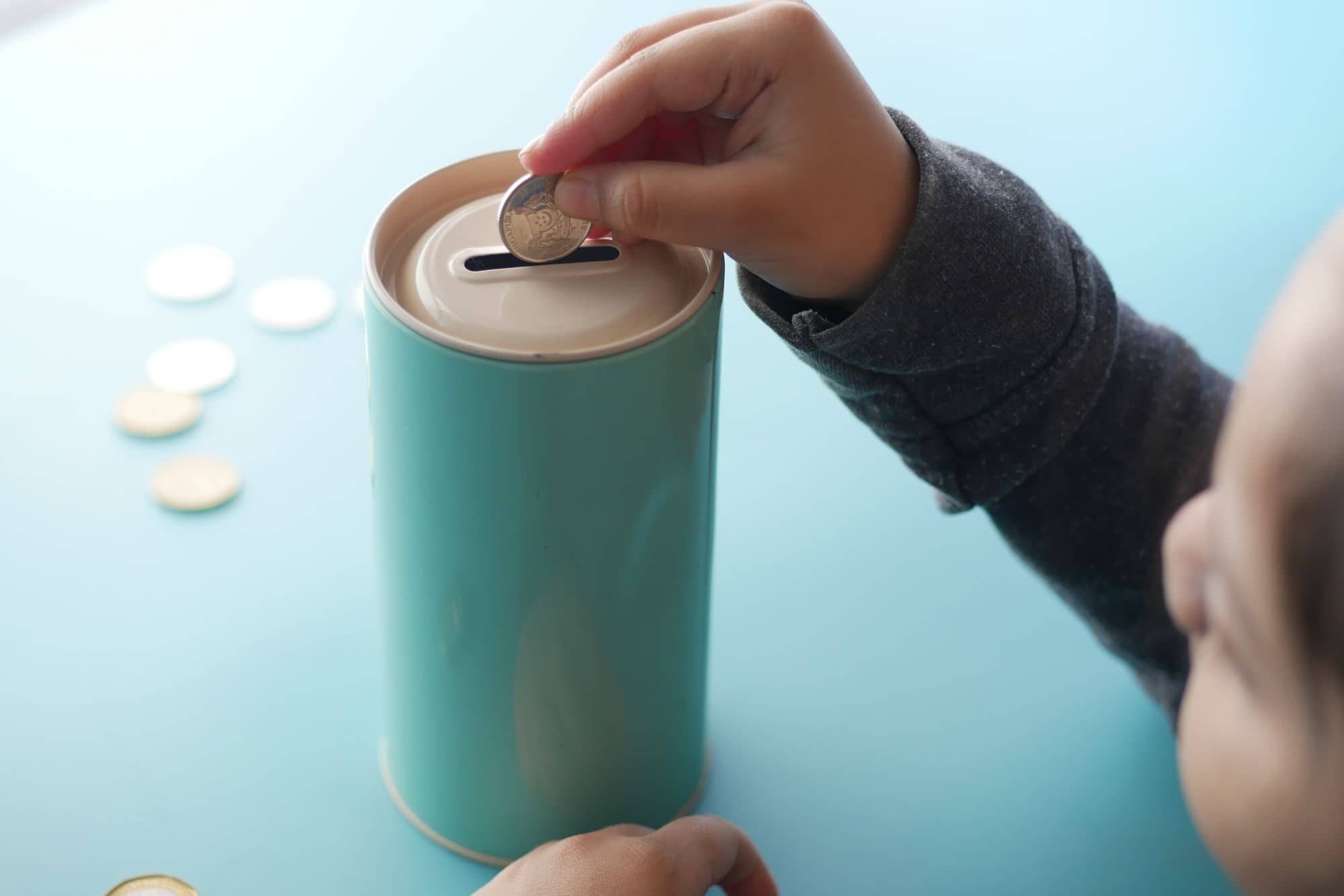 A child's hand drops a coin into a teal coin bank