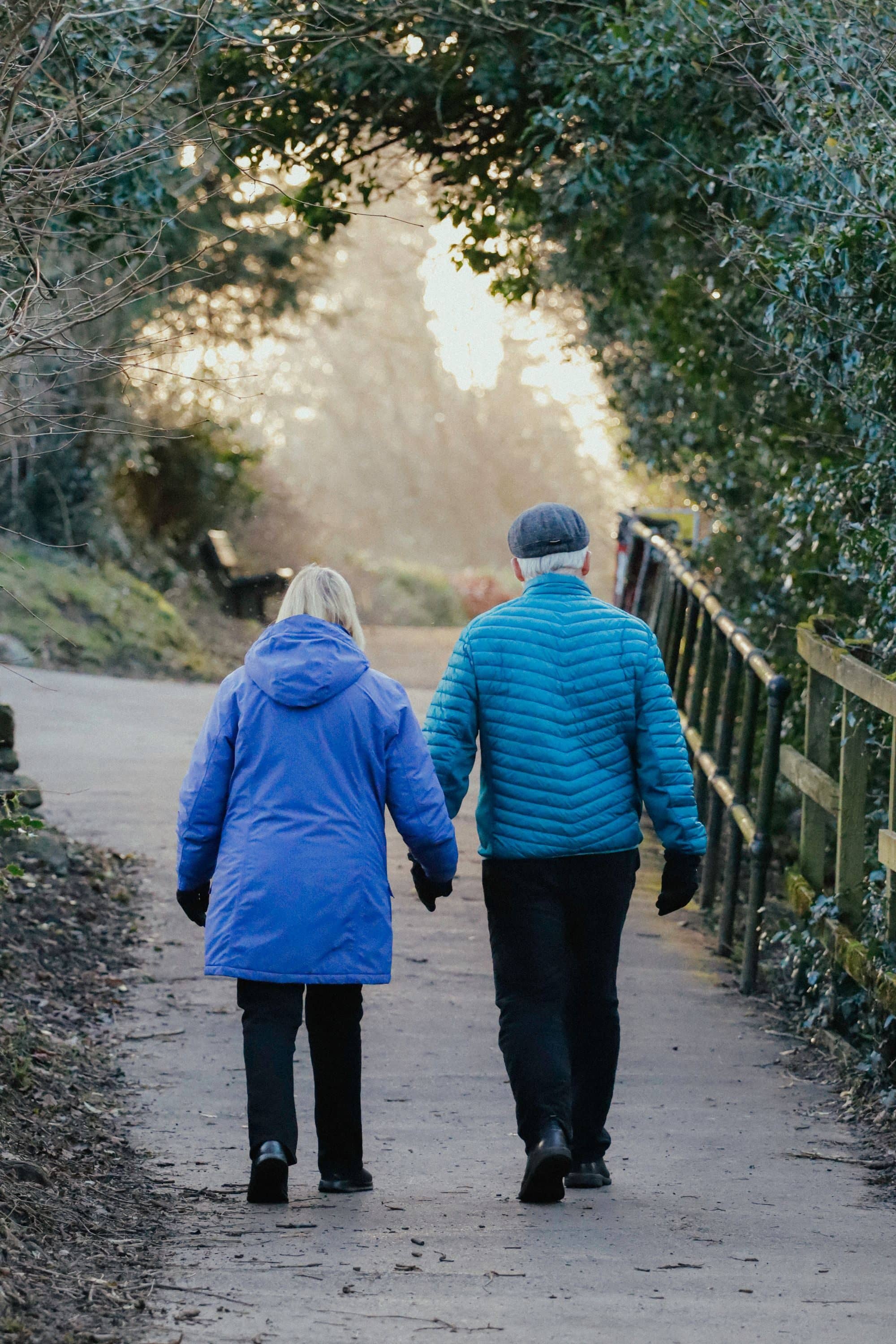 An older couple in winter jackets walks along a wooded path