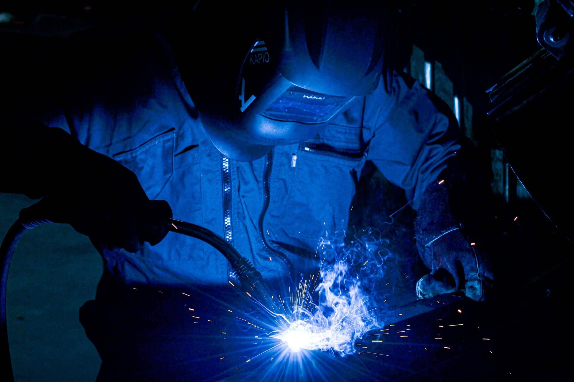 A welder in a protective helmet and blue coveralls welds steel as sparks fly