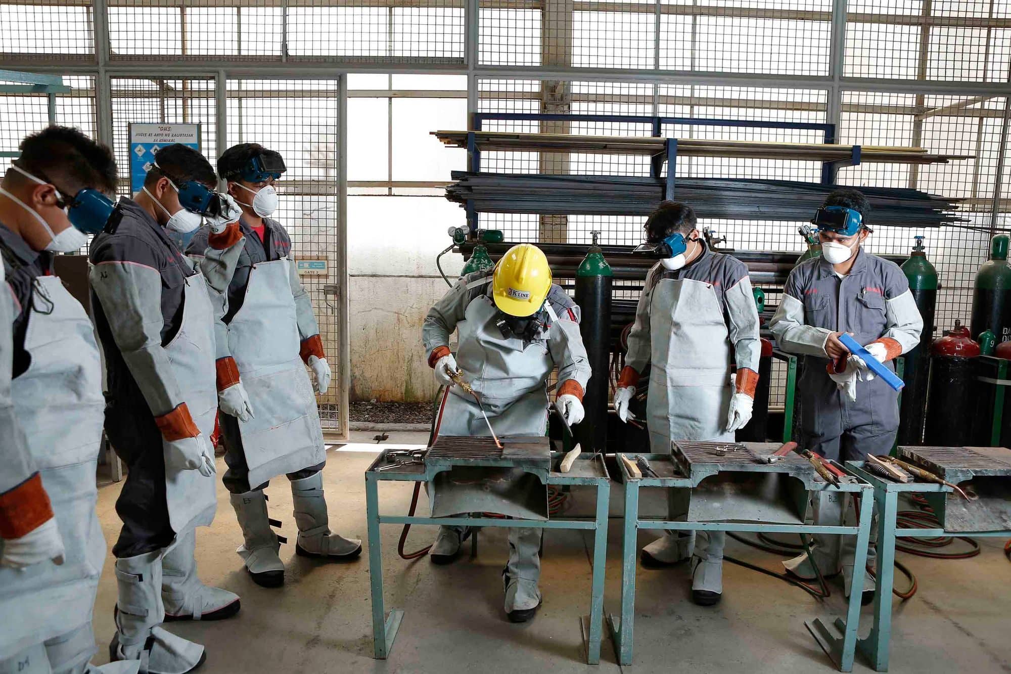 Welding students in leather aprons and safety glasses at work benches in a training shop