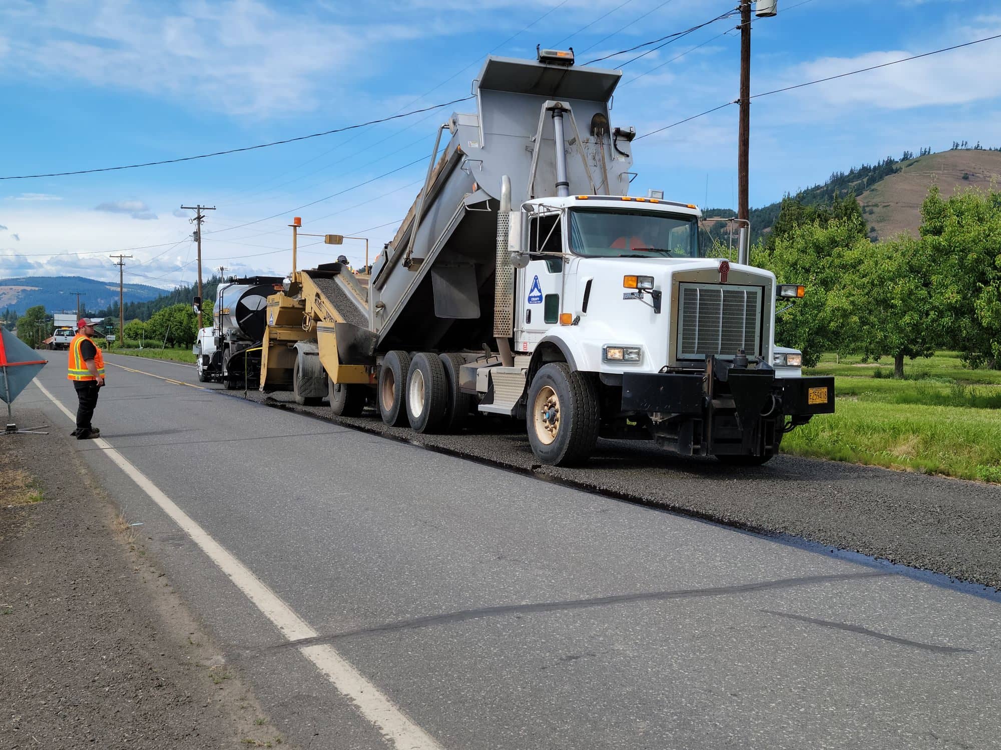 A dump truck feeds hot asphalt into a paver on a two-lane road while a flagger directs traffic