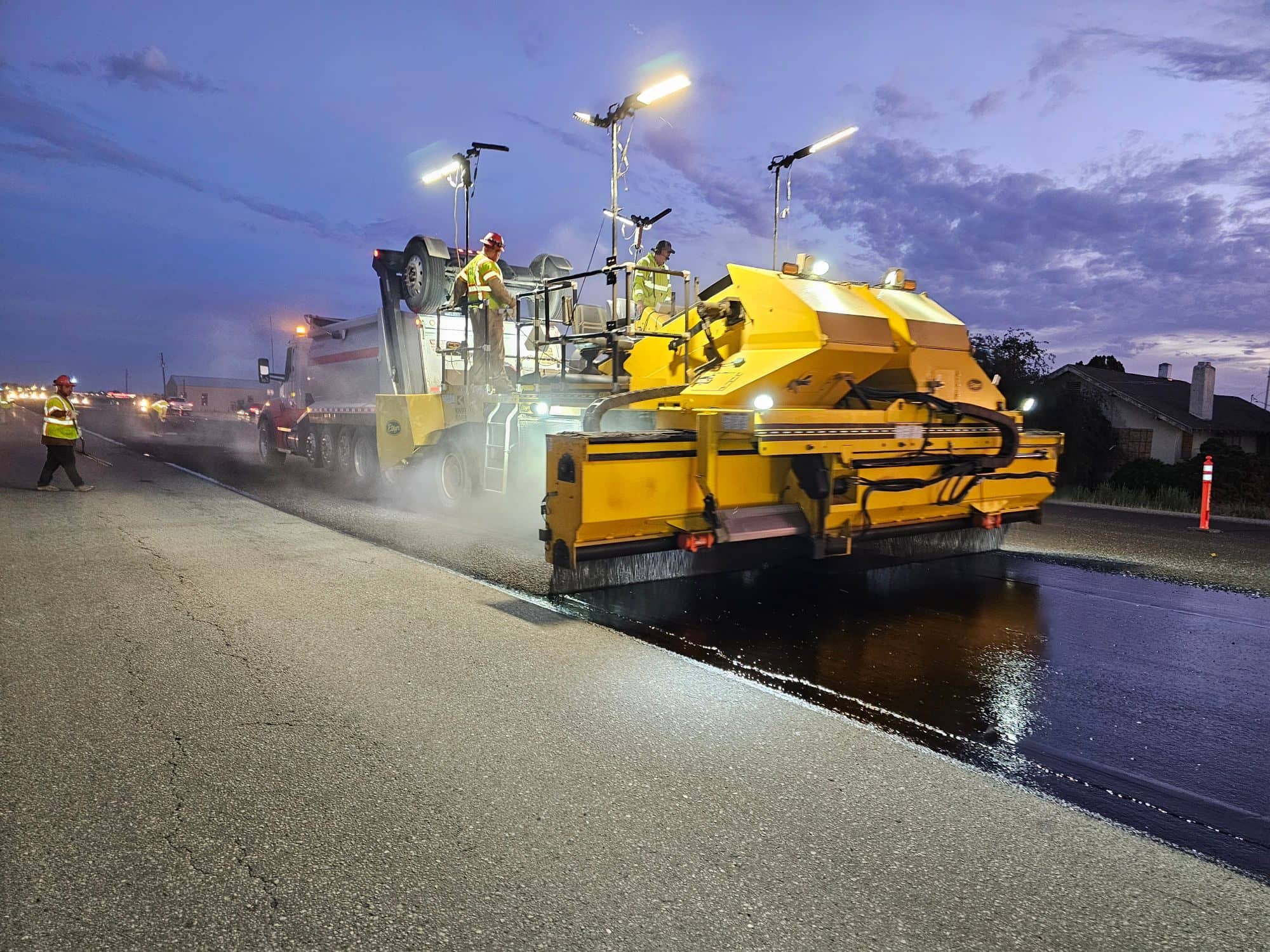 An asphalt paver lays a fresh lane at dusk under work lights while crew members walk alongside
