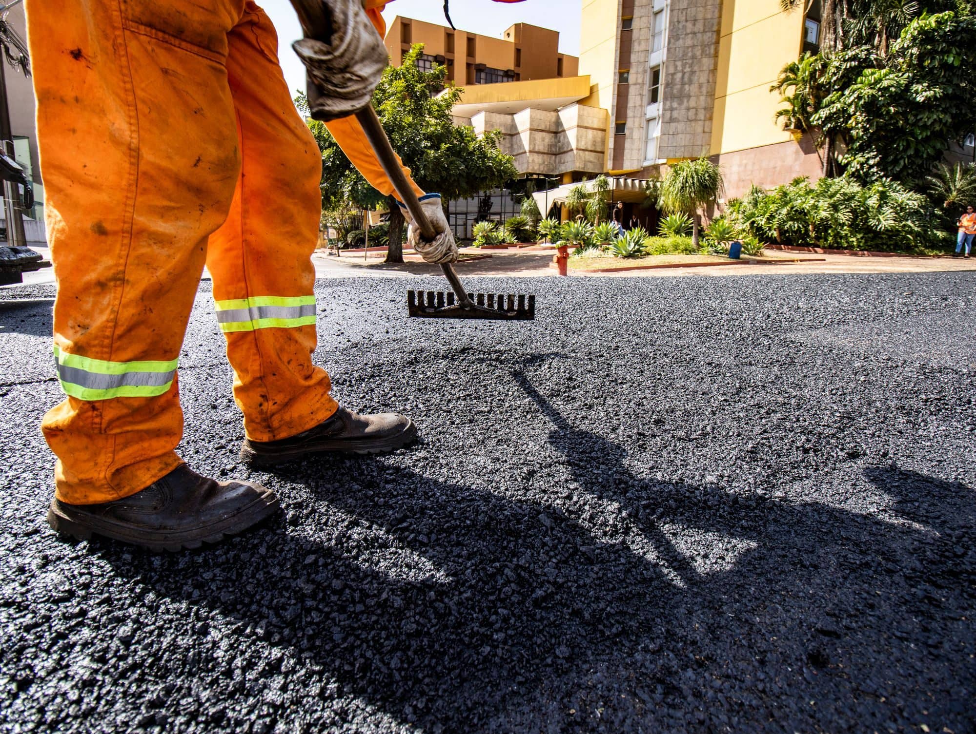 A worker in orange high-visibility pants rakes fresh hot asphalt