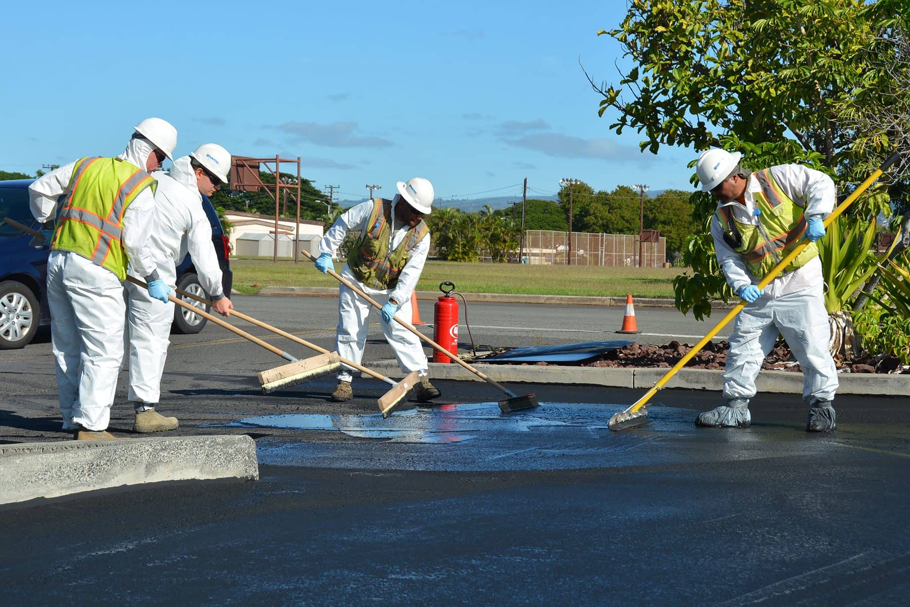 Four workers in white coveralls, hard hats and safety vests spread fresh black sealcoat across a parking lot with squeegees