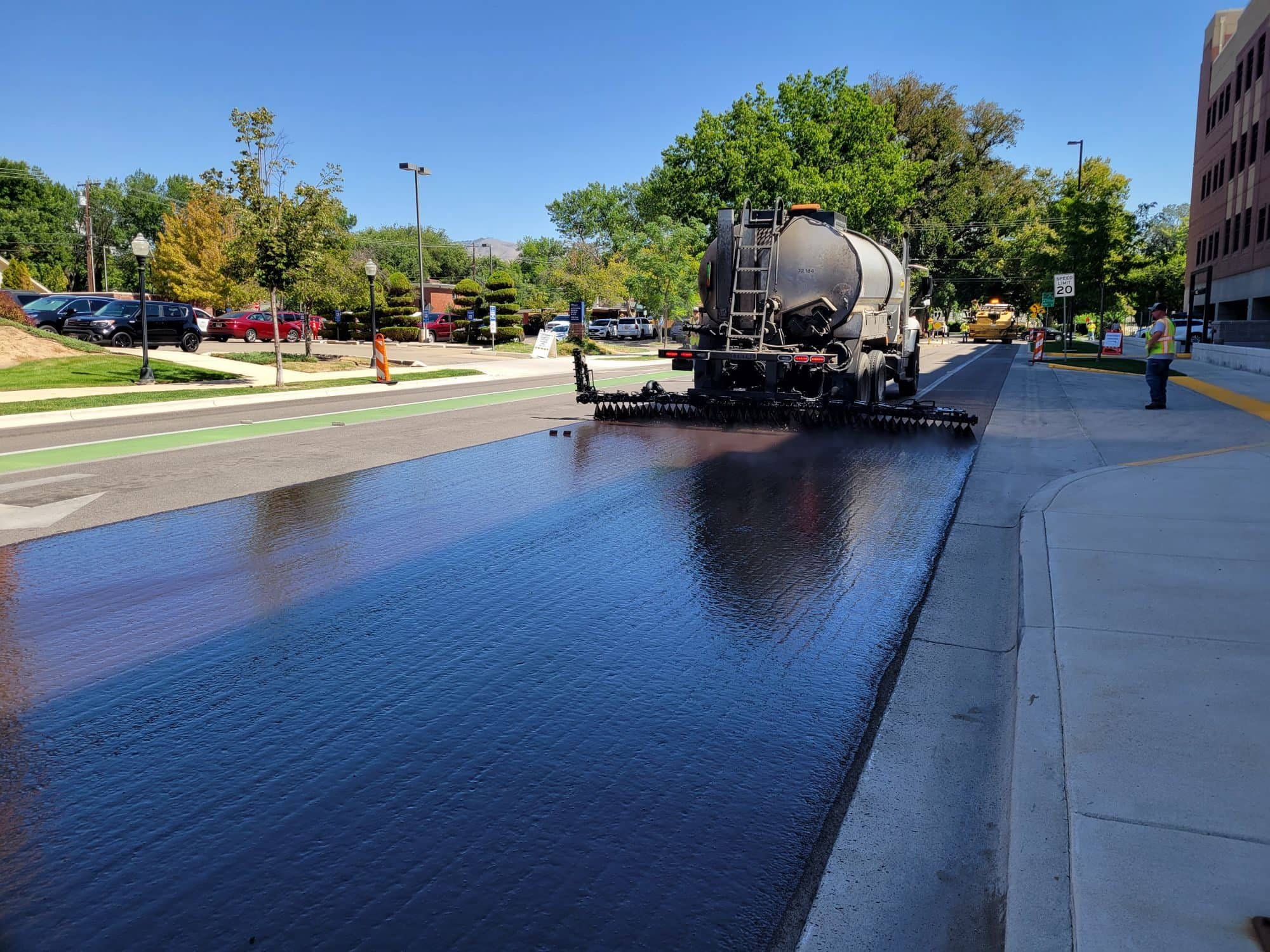 A tanker truck sprays a glossy black seal coat across a city street lane