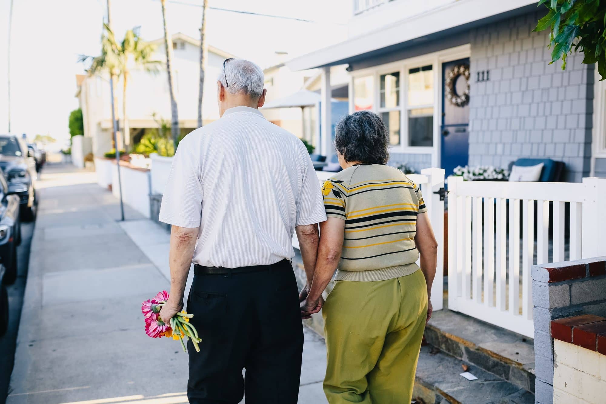 An older couple walks hand in hand down a sidewalk past houses, one holding flowers