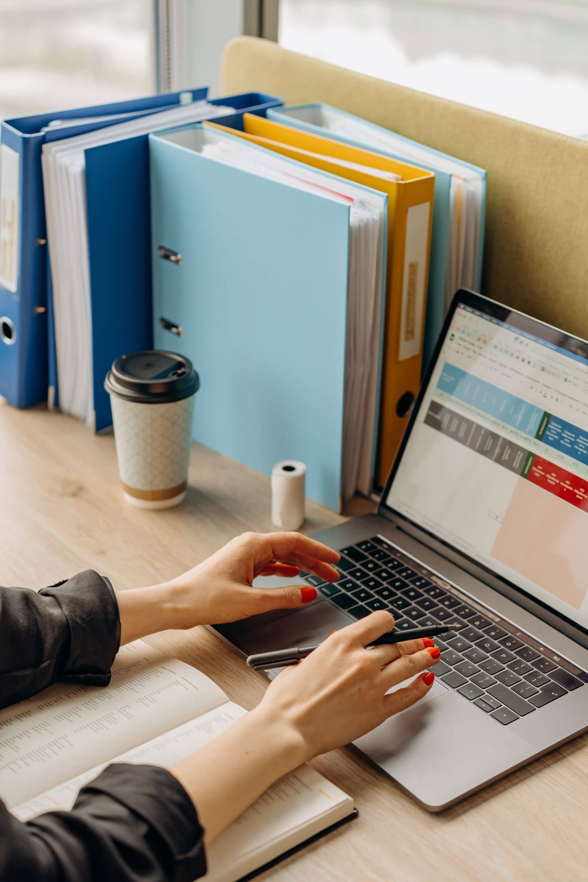 A person types on a laptop showing a spreadsheet beside a shelf of binders