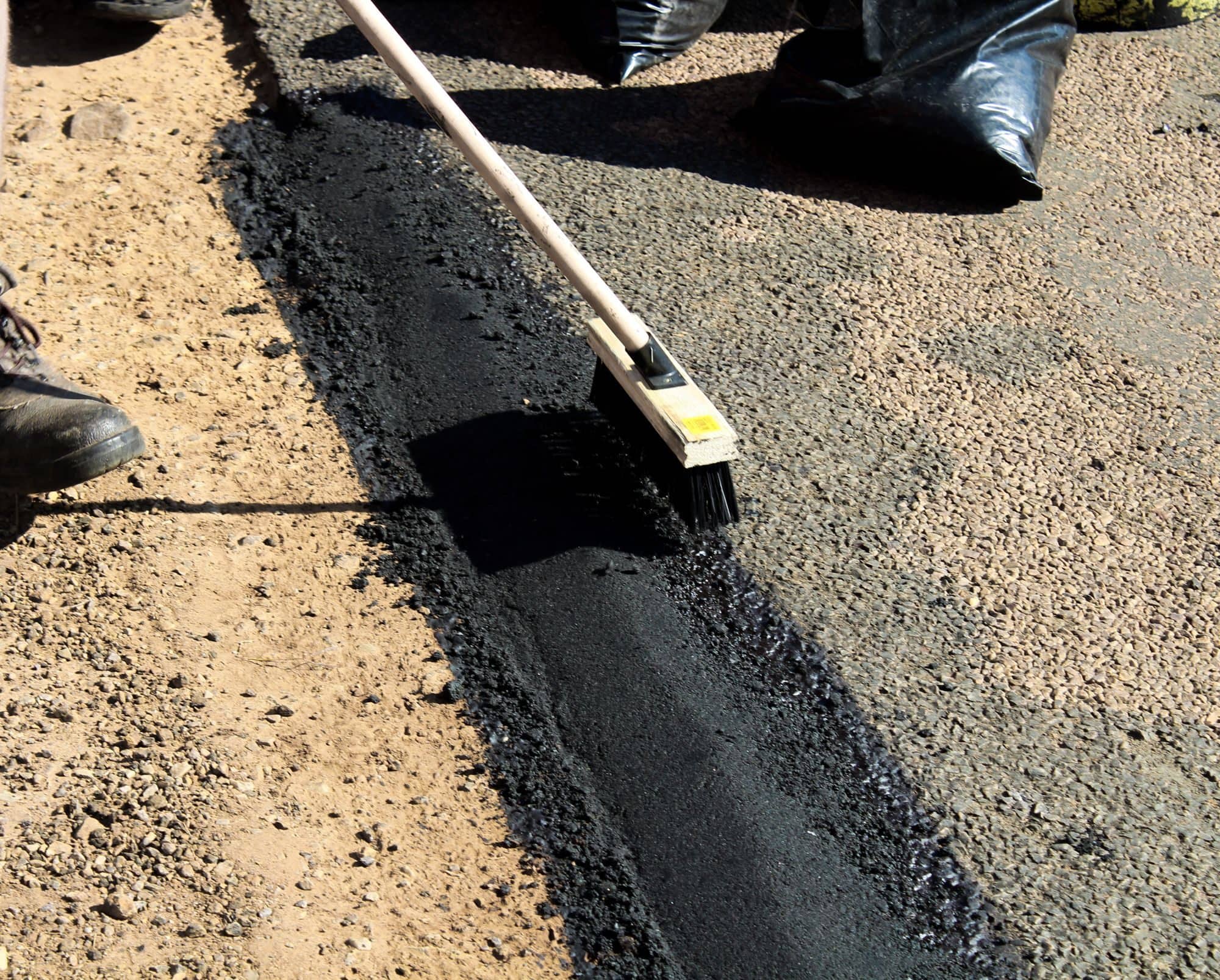 A long-handled brush spreads black sealant along a seam in the pavement