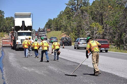 A road crew in safety vests and hard hats sweeps a freshly sealed highway lane with push brooms, a dump truck behind them
