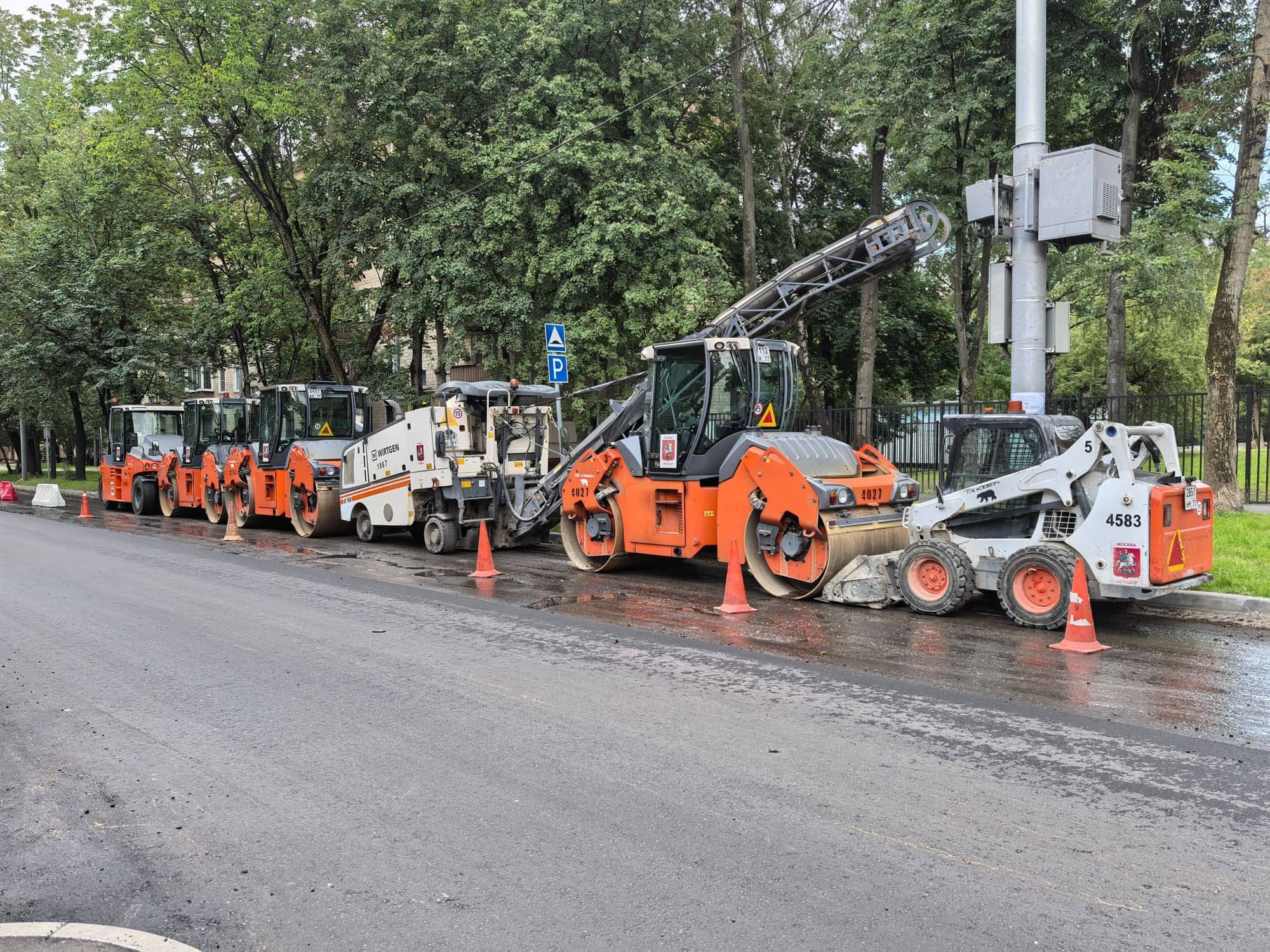 Rollers, a skid steer and other paving equipment lined up along a city street behind traffic cones