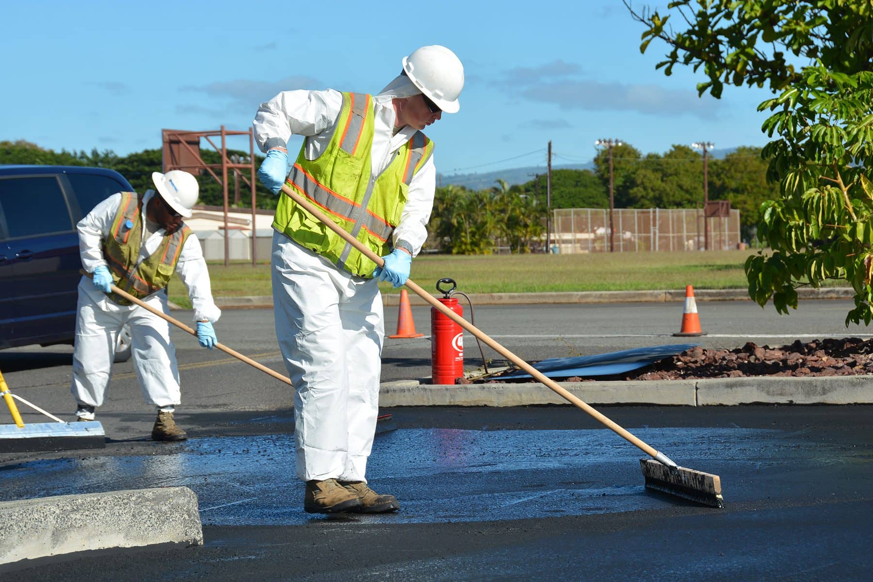Two workers in white coveralls and hard hats squeegee sealcoat along a parking lot curb