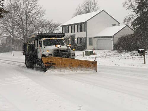 A plow truck pushes snow off a residential street in front of a snow-covered house
